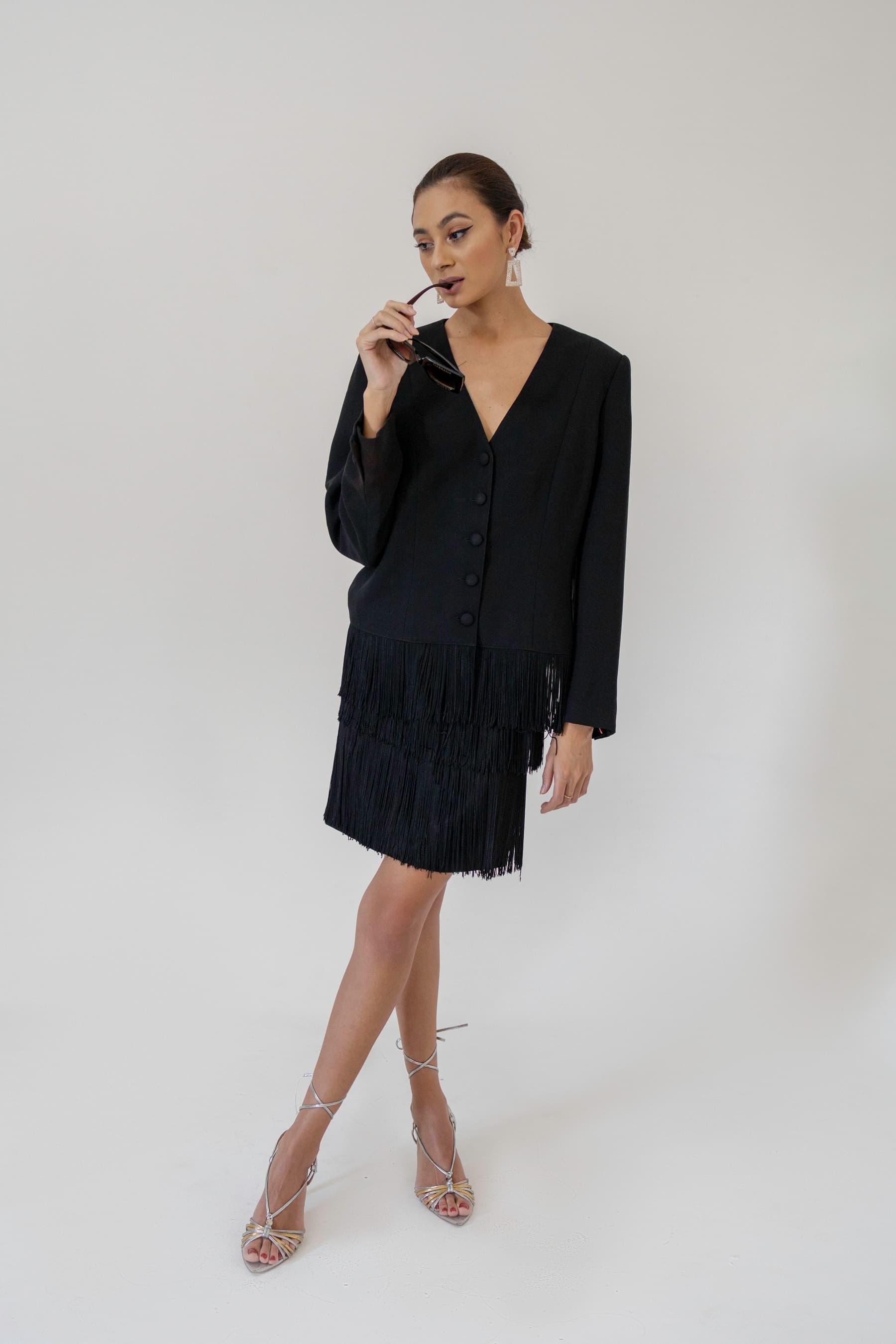 Julia Jones modeling black fringed tailoring, sunglasses, and metallic heels in a white studio